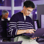 Um homem de óculos trabalha em um laptop em um escritório moderno com vários monitores exibindo códigos e simbolizando o que é defesa cibernética.