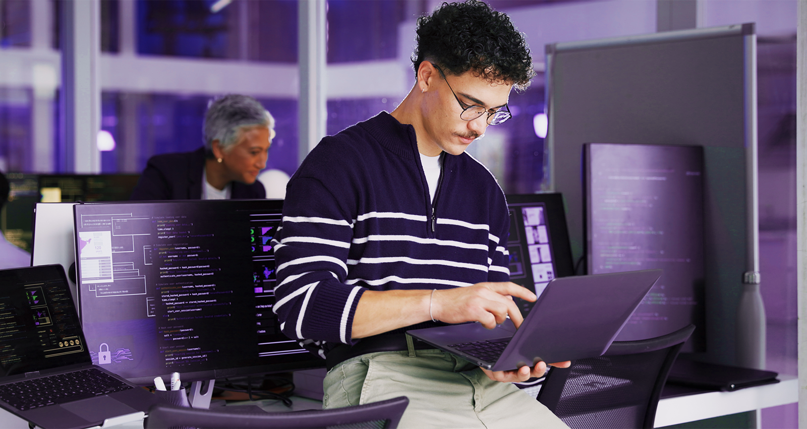 Um homem de óculos trabalha em um laptop em um escritório moderno com vários monitores exibindo códigos e simbolizando o que é defesa cibernética.